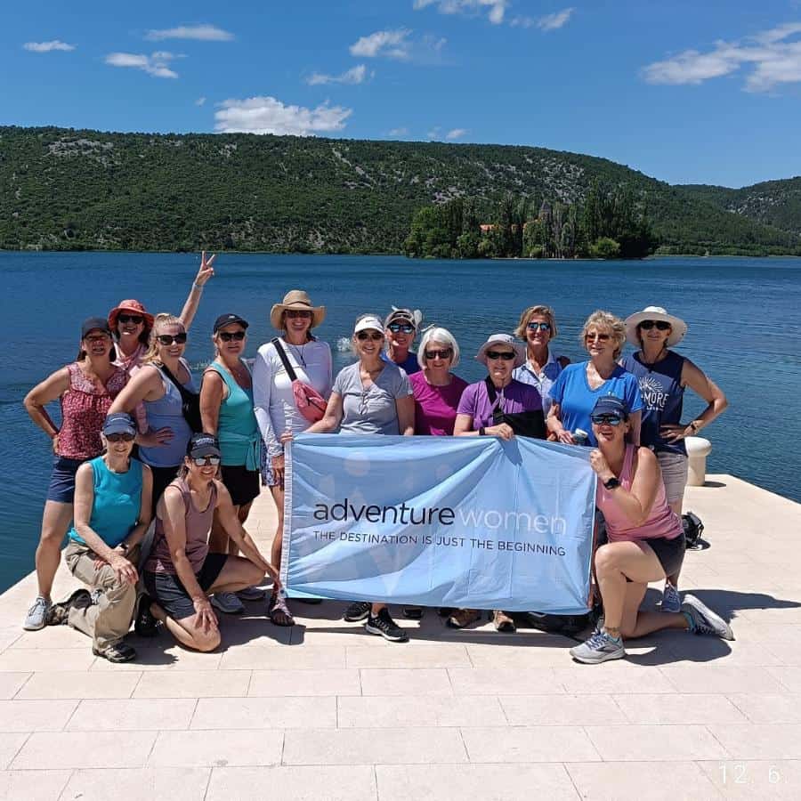 Fifteen middle-aged women stand on front of the turquoise sea. A few of them are holding a flag that says "Adventure Women, The Destination is Just the Beginning".