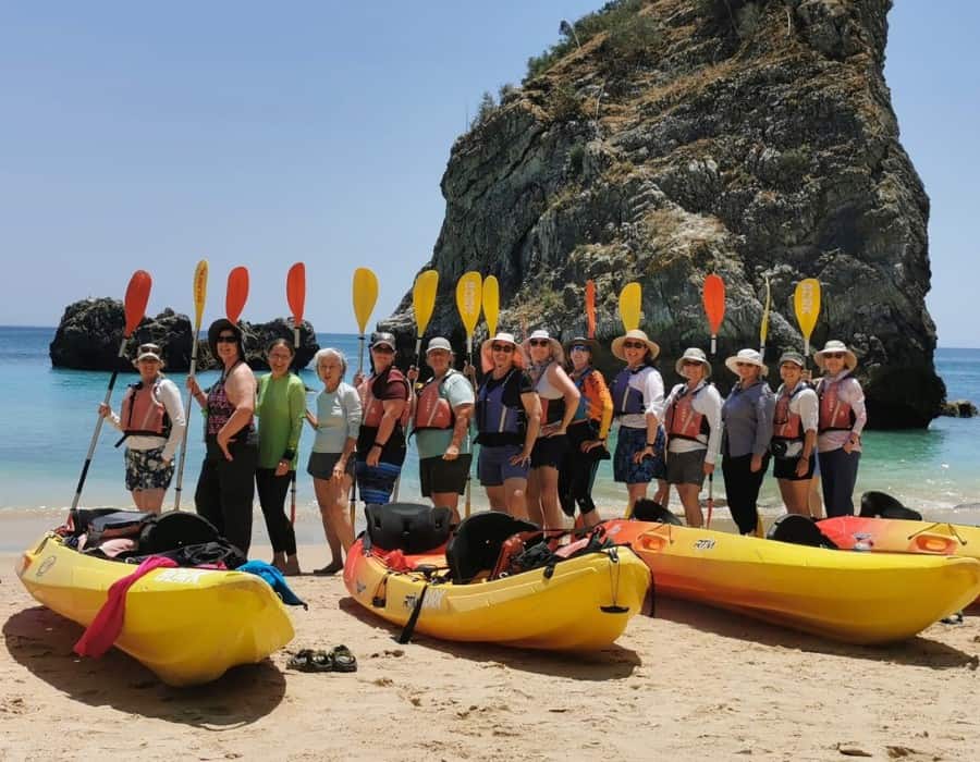 Fourteen women stand next to kayaks, holding paddles in the air, on a Portugal beach. They are on a group tour with Adventures in Good Company.