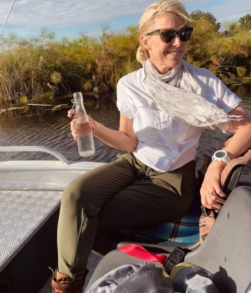 Susan Heinrich sits in a boat on an African safari dressed in green pants, a white shirt and a scarf and sunglasses. She is in the Okavango Delta with water and papyrus plants beyond.