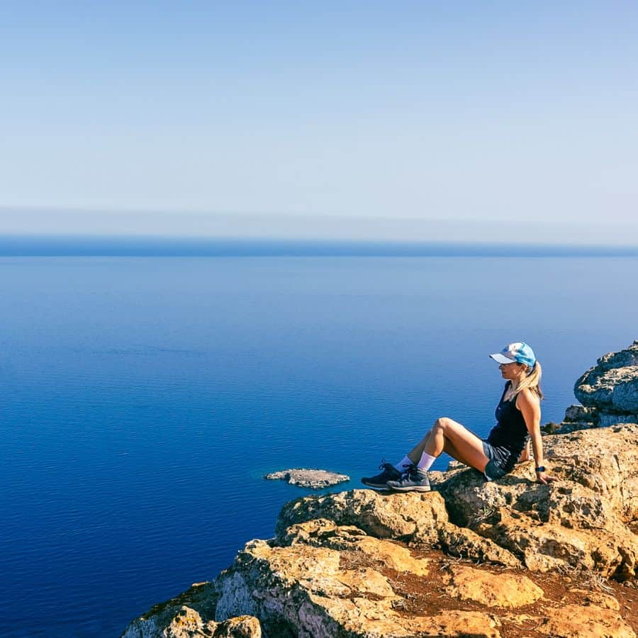 A woman sits on a rocky ledge looking out over the Mediterranean Sea far below
