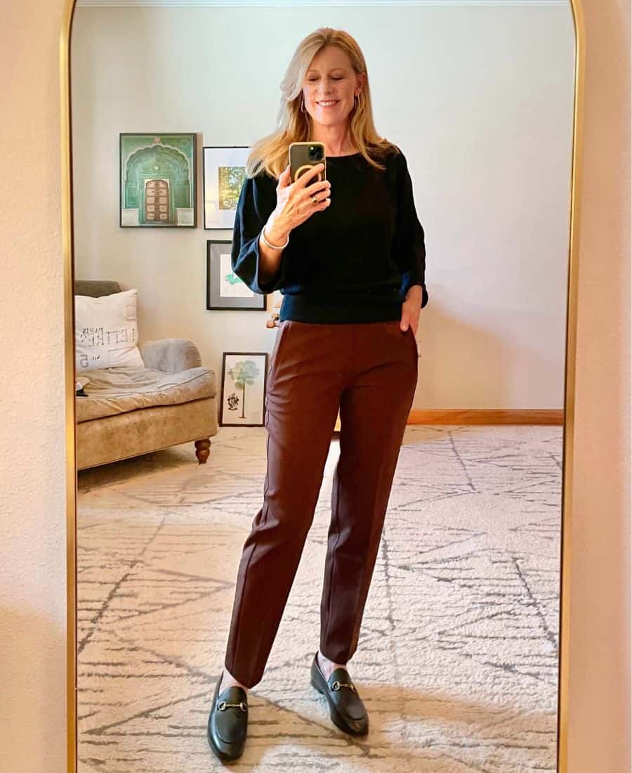 A midlife woman is reflected in a mirror wearing brown pants and a black sweater and loafers