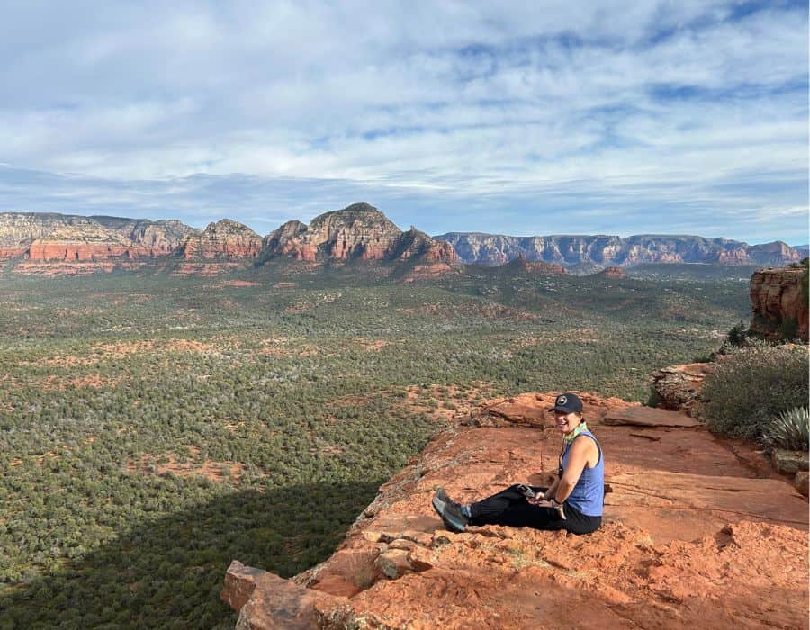 A woman sits on a red rock cliff overlooking a vast green and red canyon.