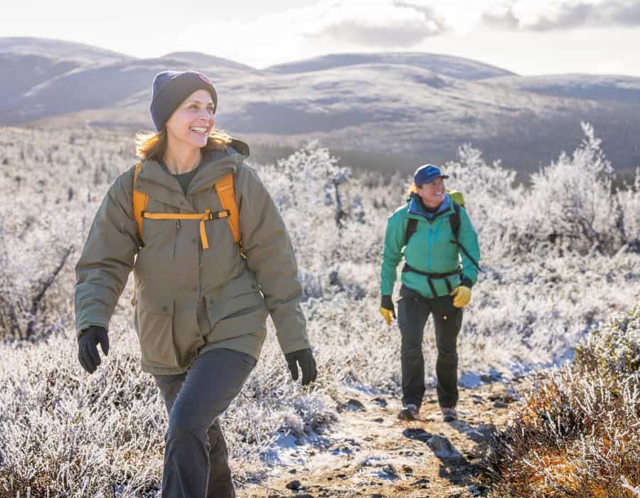 Two women are hiking in an Arctic Landscape, with snow covered trees and hills in the background.
