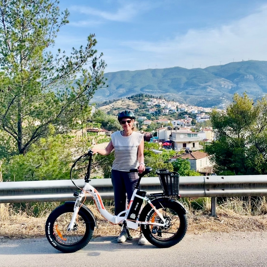Susan Heinrich with an e-bike on the Greek island of Poros, a town on a hill in the distance