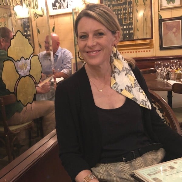 Susan Heinrich sits in a traditional Paris bistro.l She is wearing a black top and a scarf in her hair.