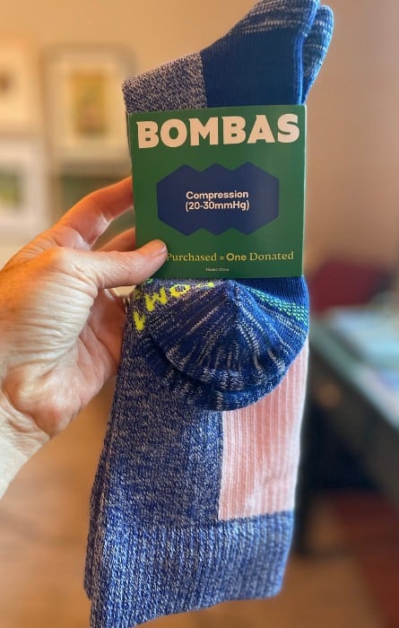 A hand holds a pair of compression socks for travel. They are blue and pink and folded inside a cardboard sleeve with the brand name Bombas on it.