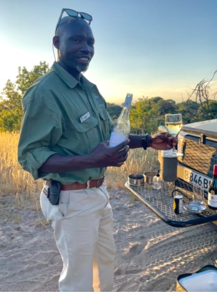 Ollie, a guide at Leroo La Tau safari camp pours a glass of wine for a guest at sundowner time in Botswana. The sun sets beyond him and the edge of a safari jeep is also visible with drinks set out in it.