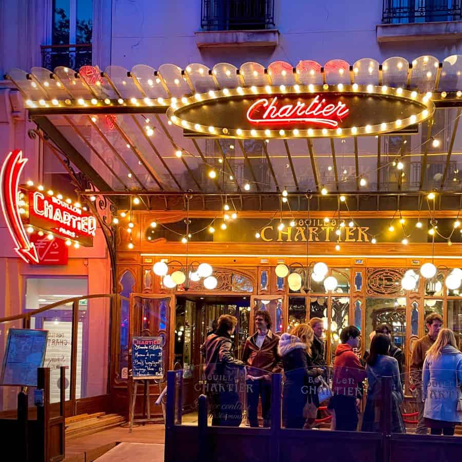 The front exterior of Bouillon Chartier at night with a group of people waiting outside. The outside is lit with art nouveau style lights and two bright signs that say Chartier