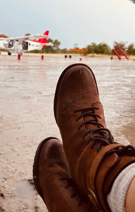 Feet in African safari boots with a bush plane beyond in a muddy landing strip in Africa.