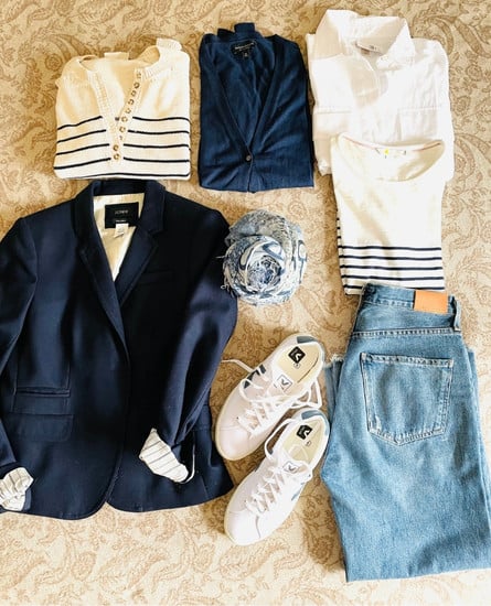 A capsule wardrobe for spring starts with a neutral palette of navy and white. Pictured are a flat lay of navy blazer, denim jeans, blue and white scarf, white shirt, navy cotton cardigan sweater, striped sweater and striped mariniere tee shirt.