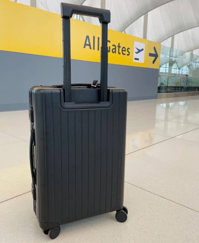 A black carry-on roller bag with the handle extended, is visible from the back. Beyond it is a bright yellow sign that says All Gates with a graphic of an airplane and an arrow.