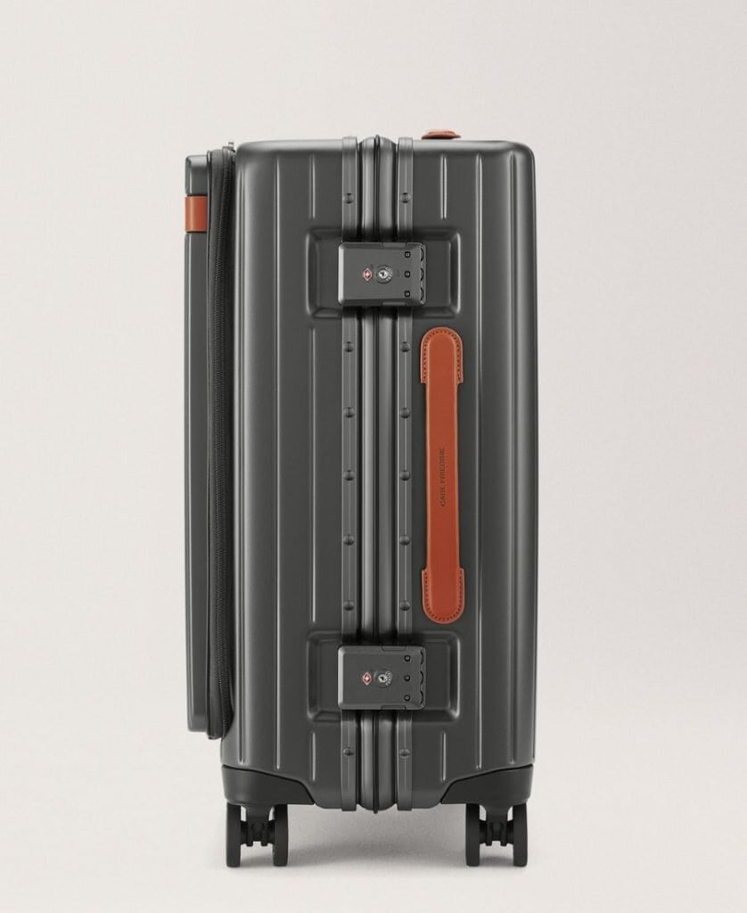 A carry-on roller bag is pictured from the side view. It is dark grey with brown leather trim and has four wheels. It also has a front pocket with a zipper. It has a plain background beyond it.