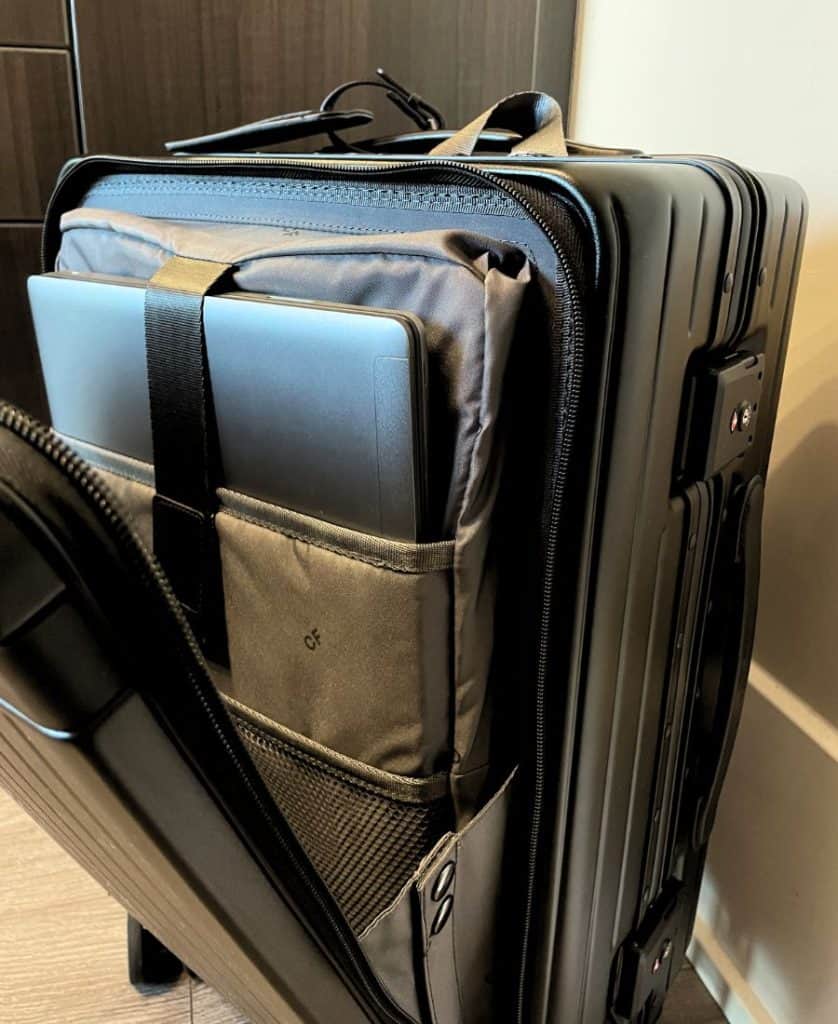 A black carry-on roller bag is shown with the front pocket zipped open. Inside are multiple pockets for a laptop and other items.