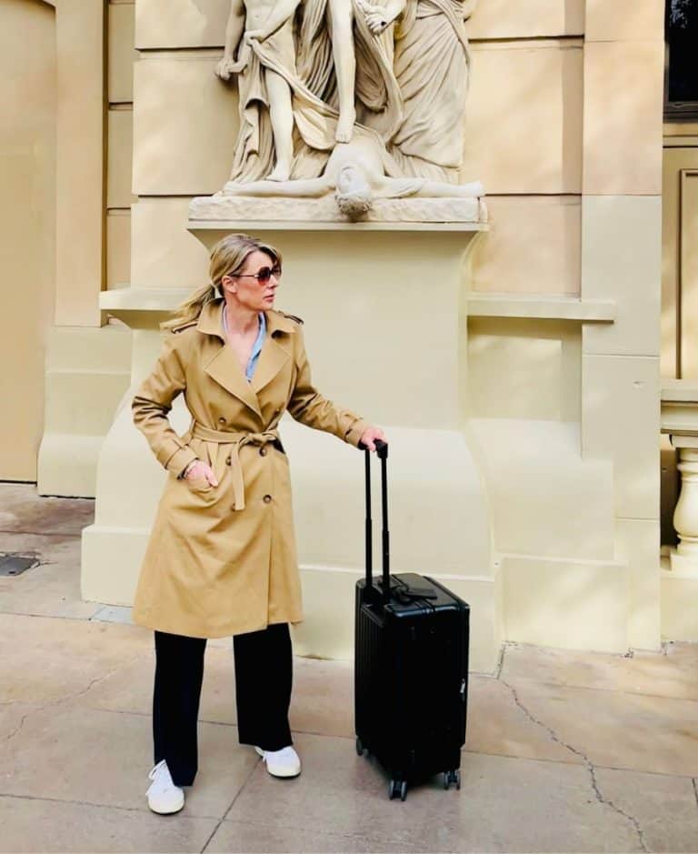 A blonde woman wearing black pants and a beige trench coat and sunglasses stands in front of what looks like a European building. She is holding the extended handle of a black carry-on roller bag.