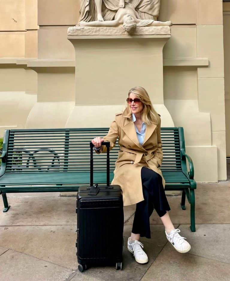 A blonde woman wearing black pants and a beige trench coat and sunglasses sits on a green bench with a statue partially visible above her. She is holding the extended handle of a black carry-on roller bag.