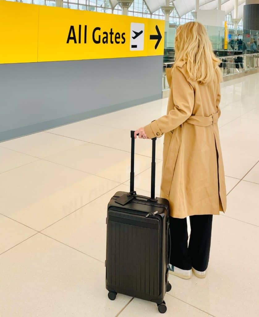 A woman is visible from the back. She is blonde and wearing a beige trench coat and holding the handle of a black carry-on roller bag. She is facing a bright yellow sign that says All Gates with an arrow.