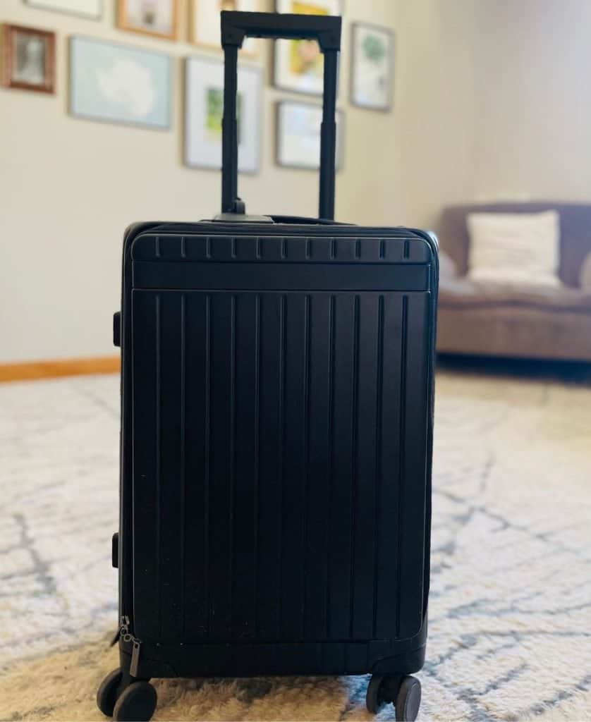 A black carry-on roller bag stands on a white and grey carpet with the handle extended. An armchair and some artwork are visible on the wall beyond.