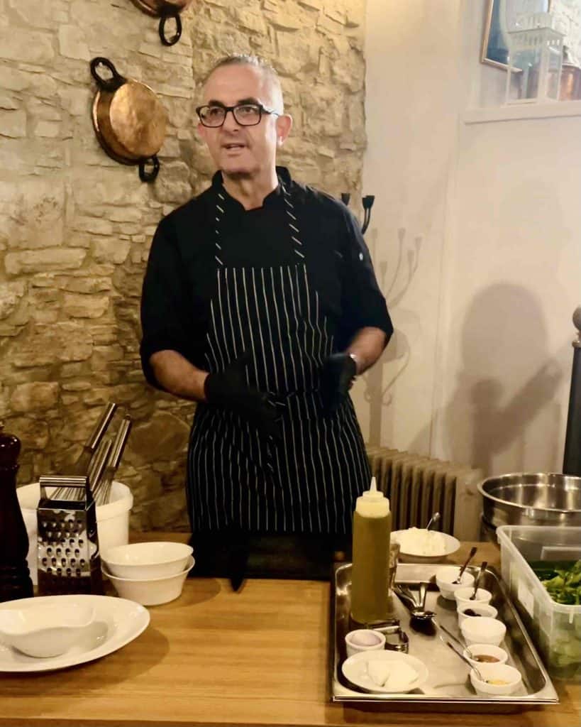 A chef in a black apron and black gloves stands behind a wooden table that is set with ingredients. Beyond him is a stone wall with two copper pots hanging on it.