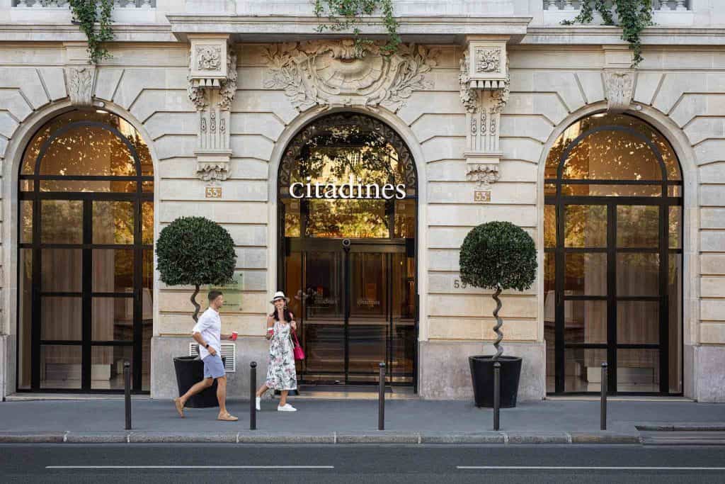 The exterior of a Paris hotel with ornate arched windows and a limestone exterior. Two people are pictured in front of the hotel.