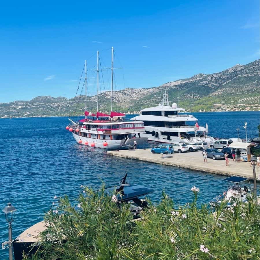 Two yachts are pictured docked on an island with a blue sea around them and a hilly landscape beyond. There are some flowering shrubs in the foreground.