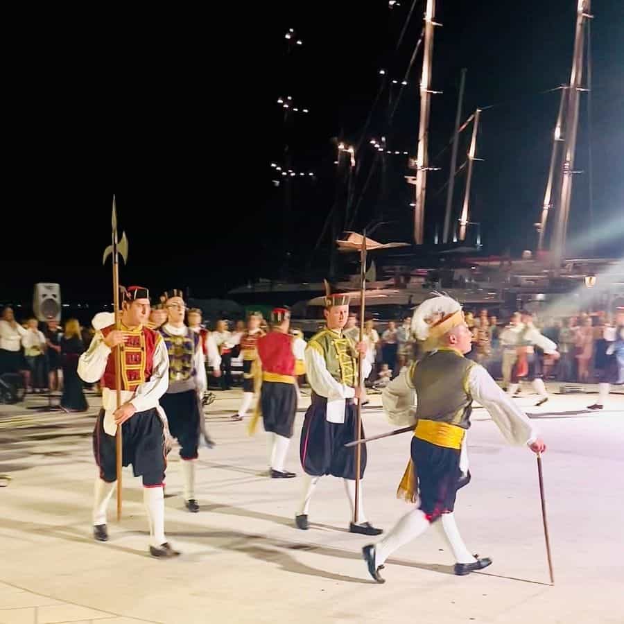 Several men are dressed in traditional outfits, dark short pants with colorful vests on top and small hats. Some are carrying long swords. The scene is well-lit but the nigh sky beyond is pitch black.