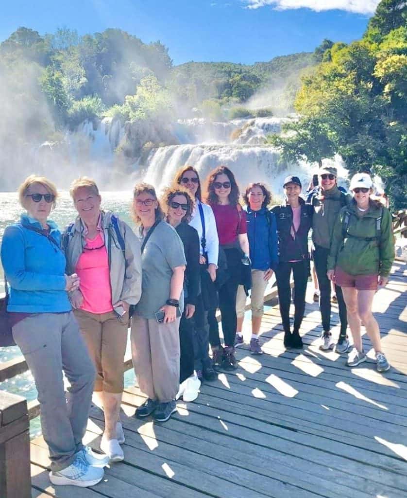 A group of women stand in front of a misty waterfall and green foliage at Krka National Park, Croatia.