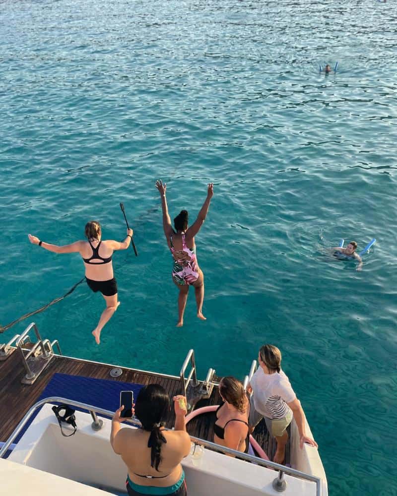 Two women jump off the back of a boat and into the turquoise sea of the Blue Lagoon of Cyprus. Other people are visible on the boat and in the water.