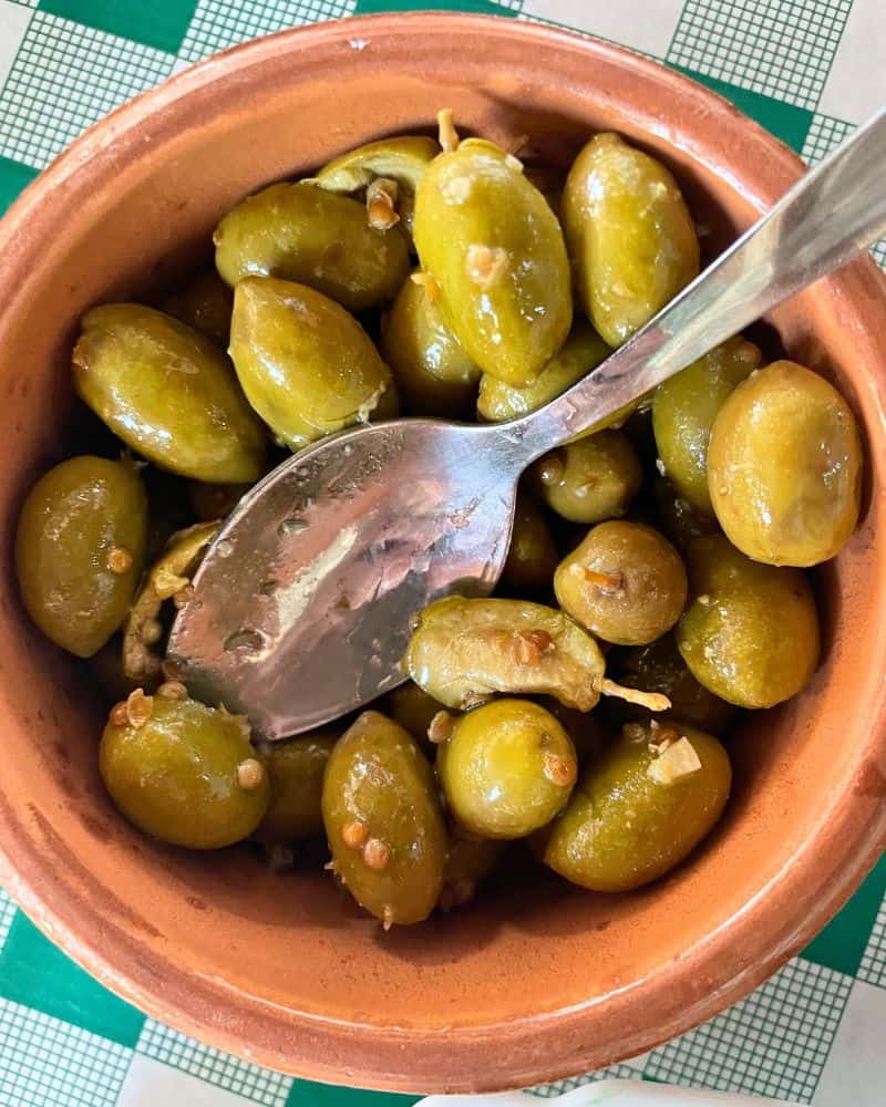 A clay bowl of green olives covered in oil and spices. A spoon is set atop the bowl.