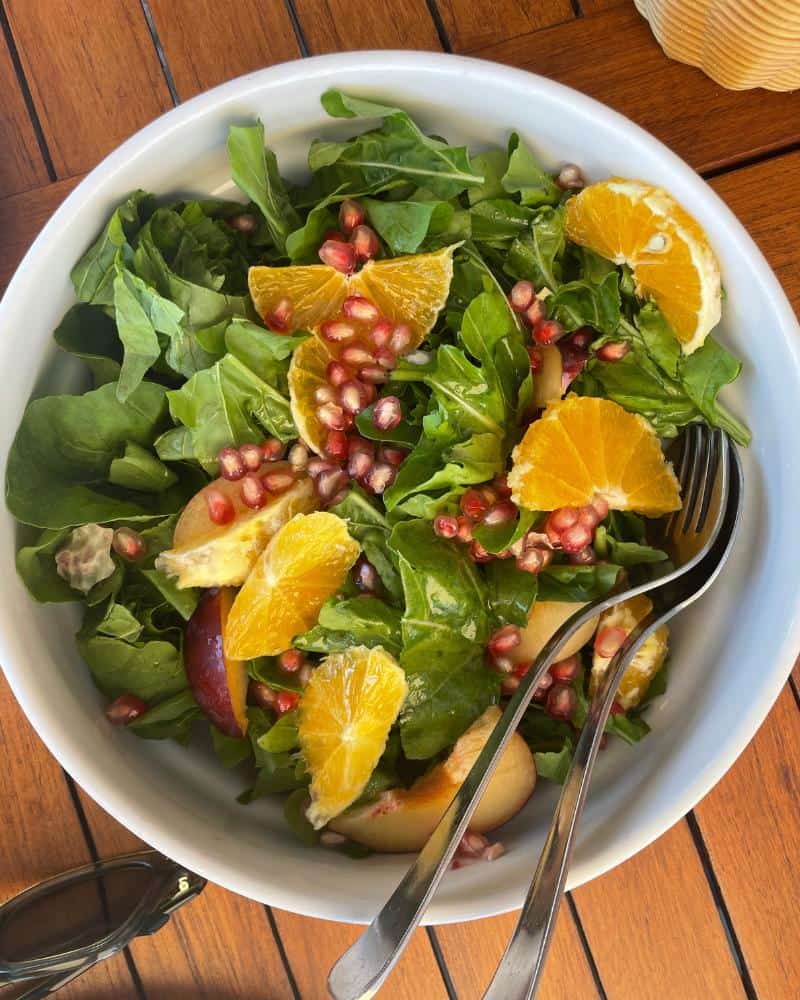 A green salad has oranges and pomegranate seeds on top. Serving utensils are set in the bowl. It's a top a wood table.