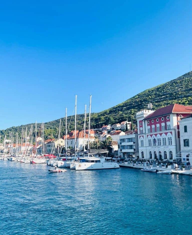 A row of sailboats are moored in the harbor of the Croatian Island of Vis. The sea is a turquoise color and the whitewashed buildings have red roofs.