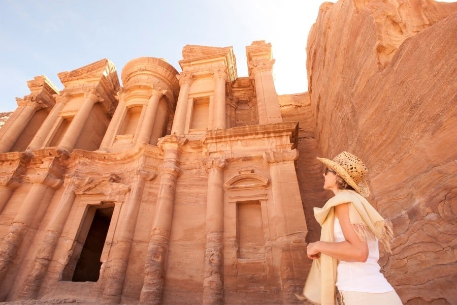 A woman traveler is pictured looking at Petra in Jordan. She is wearing a white short and shorts and a straw hat.