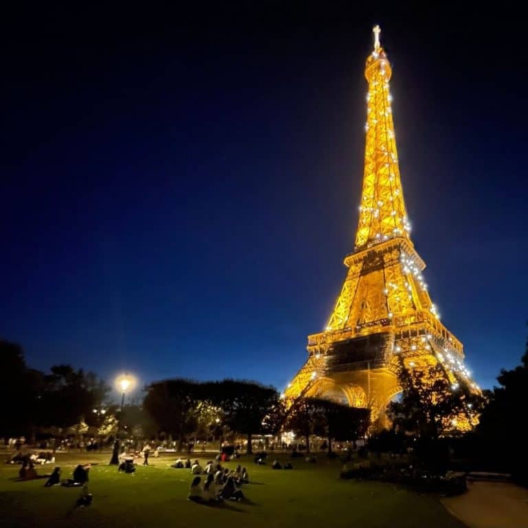 A gorgeous night time view of the Eiffel Tower from the Champ de Mars park in Paris. Visitors are spread out on the lawn in front of the tower