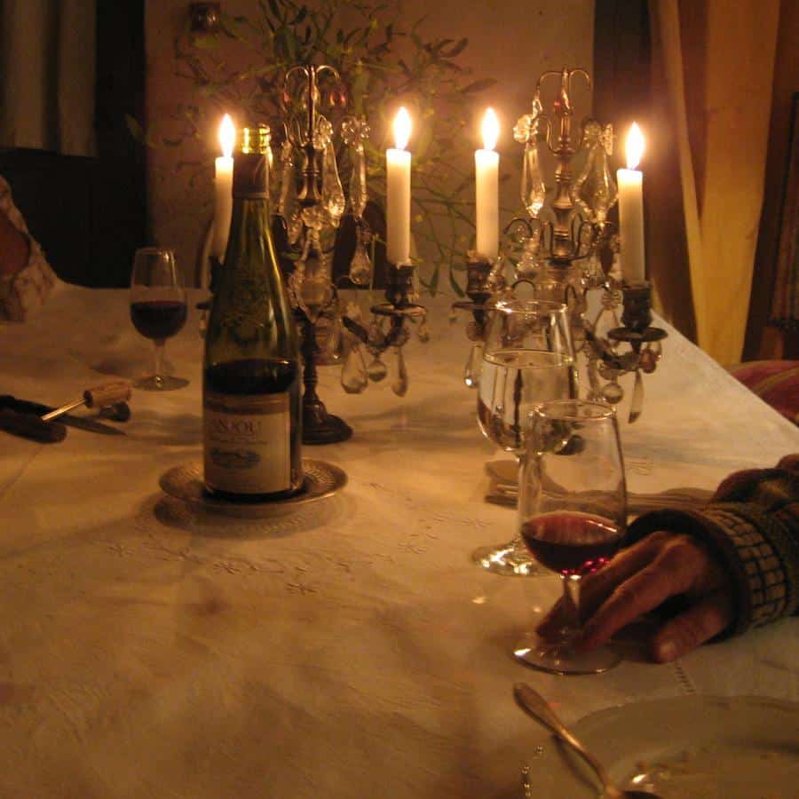A French dining table is set with candles and a bottle of wine is visible along with a hand holding a glass of red wine