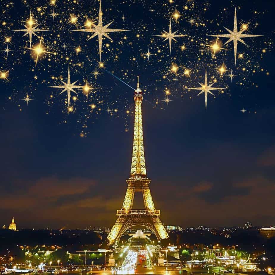 The Eiffel Tower is pictured against a dark night sky. Golden starbursts are set in the sky above it. A little of Paris is visible next to it.