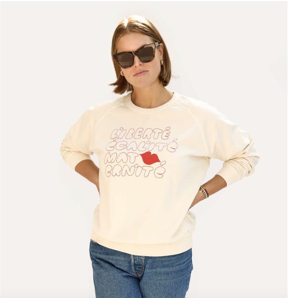 A woman wears a cream colored sweatshirt with the words "Liberté Ègalité Maternité" . It also has red lips as part of the words.