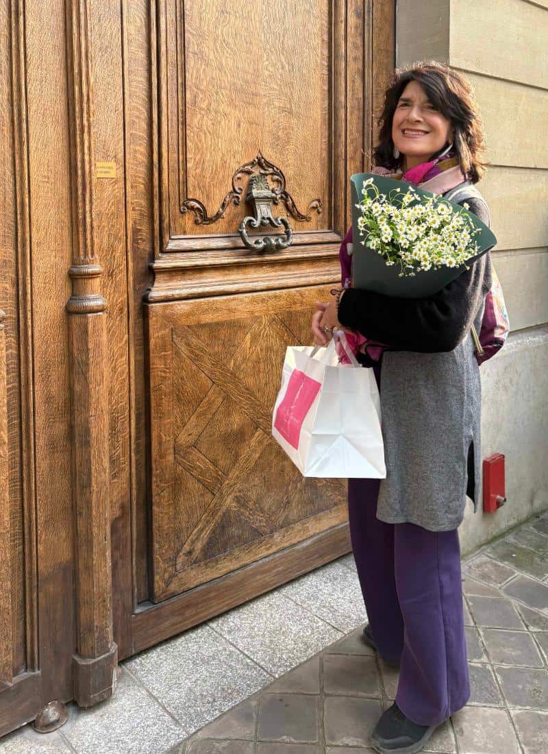 A woman stands in front of a wood door in Paris, holding a bunch of flowers