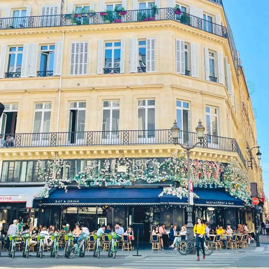 A beautiful Haussmann building in Paris with a cafe at street level with may people sitting outdoors.