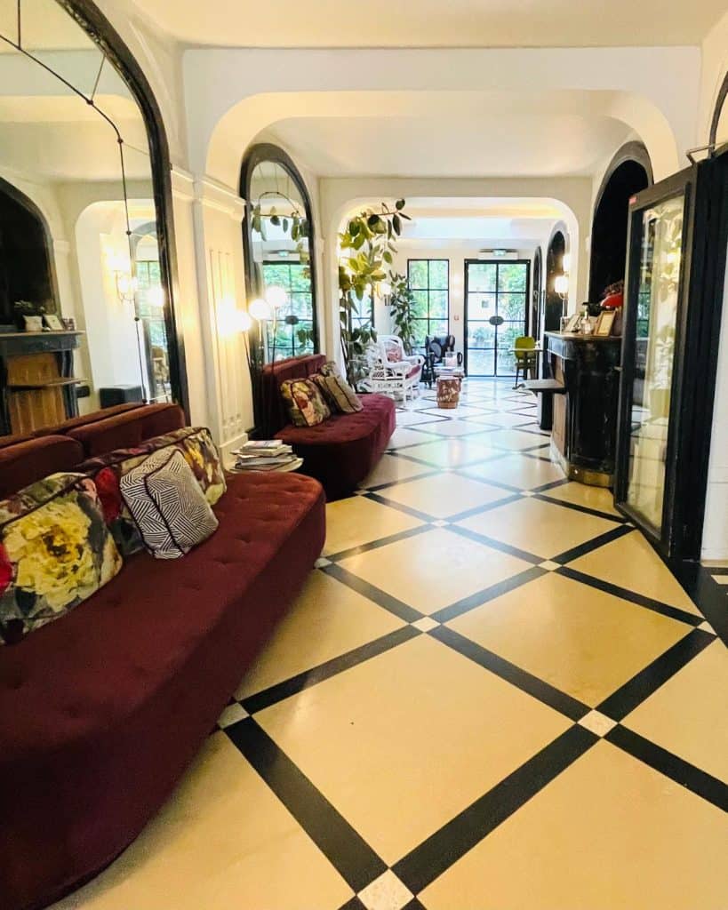 A bright hotel lobby has a diamond pattern in the stone floor and a pair of burgundy settees to one side. Doors to an outside terrace are visible at the end of the hallway. Arched mirrors reflect light back into the space making it bright and airy.