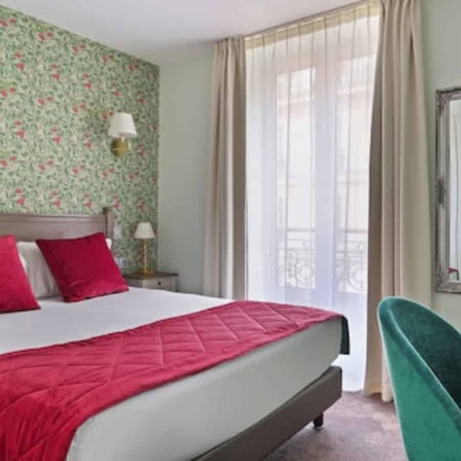 A pretty hotel room with a large floor to ceiling window, a double bed, green patterned wallpaper and red accents.