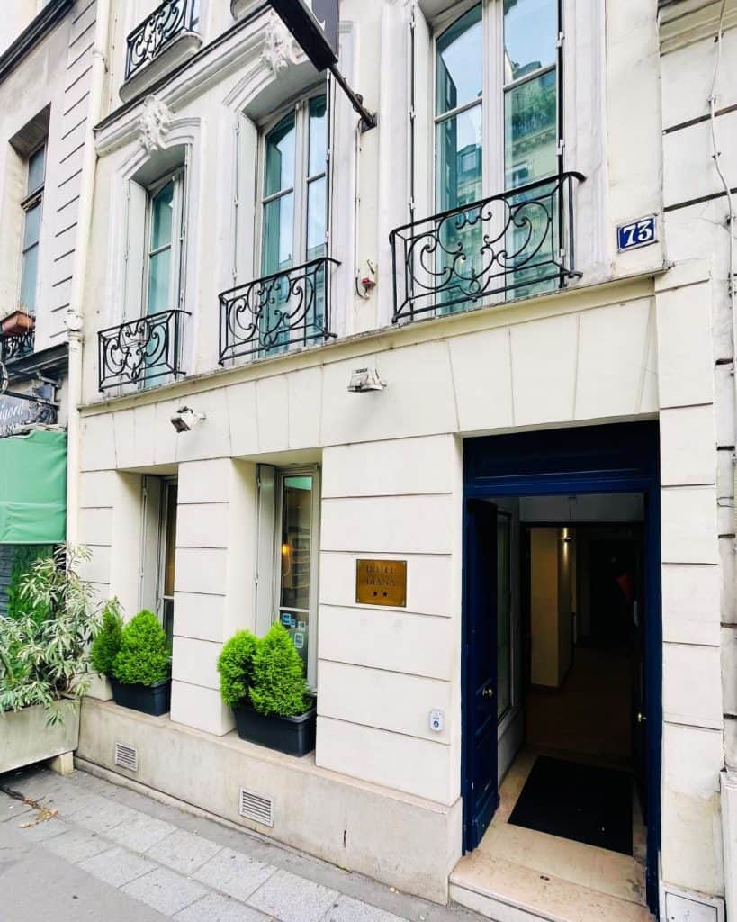 The exterior of a hotel in Paris, with an open door and window boxes with greenery. It has a brass sign with it's name and 2 stars.