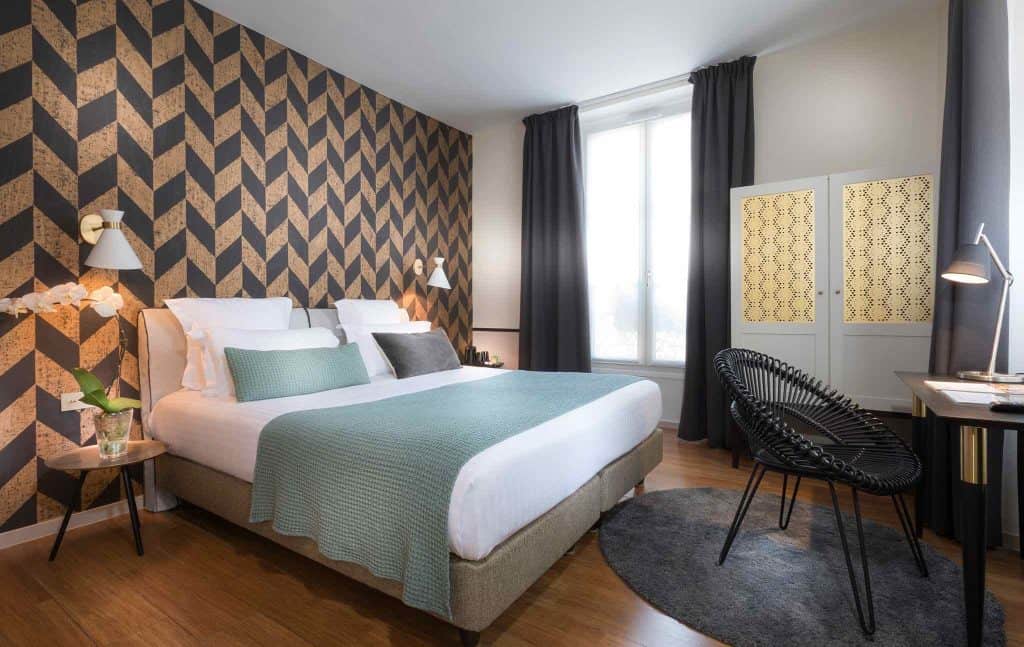 A hotel room is pictured with colorful wallpaper, a double bed with pale blue accents and a bright window.