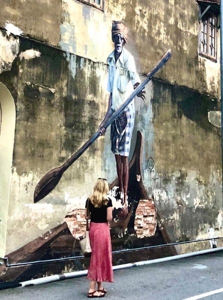Susan Heinrich stands facing a huge wall mural of an Indian Boatman in Penang Malaysia