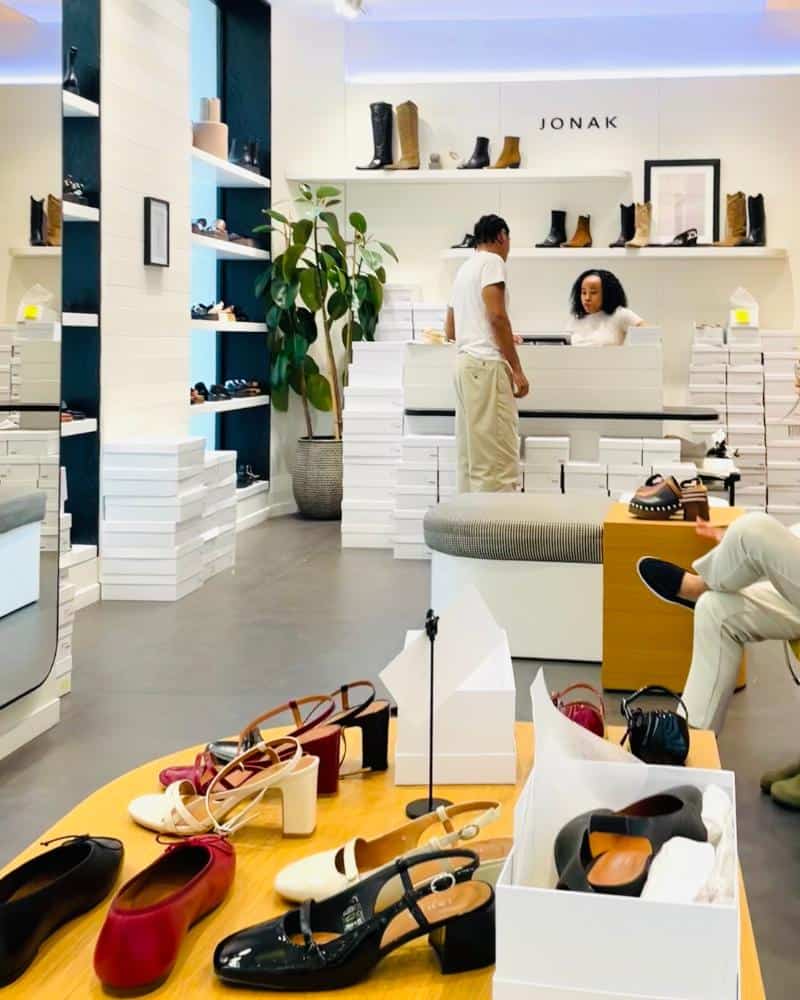 The inside of a shoe store has women's shoes on display in the foreground in colors, red, white and black. Shelves of shoes and boots and two staff members are pictured beyond.