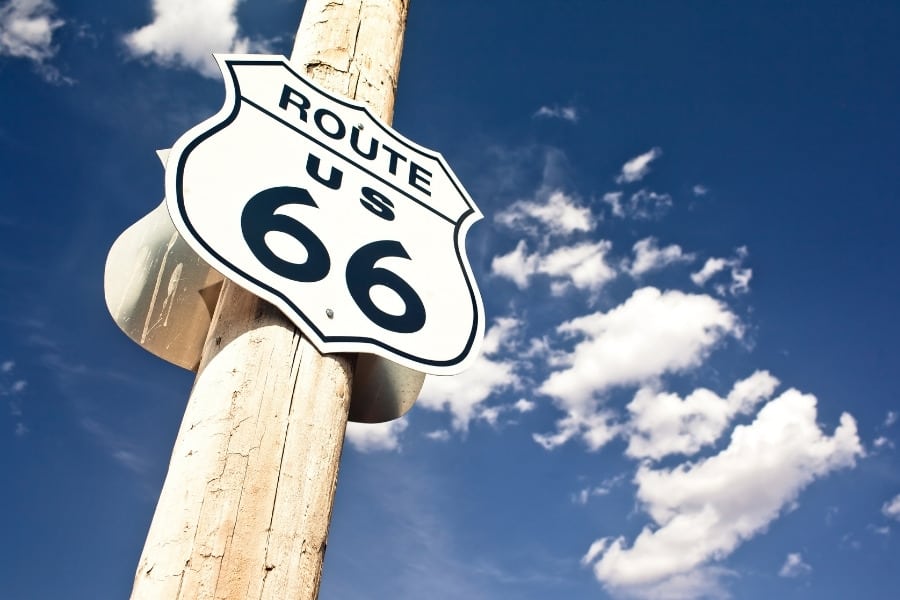A sign for Route 66 stands against a blue sky with white clouds, on a road trip between Los Angeles and Denver