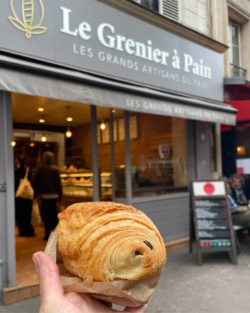A hand holds a chocolate croissant in front of Le Grenier a Pain bakery in Paris.