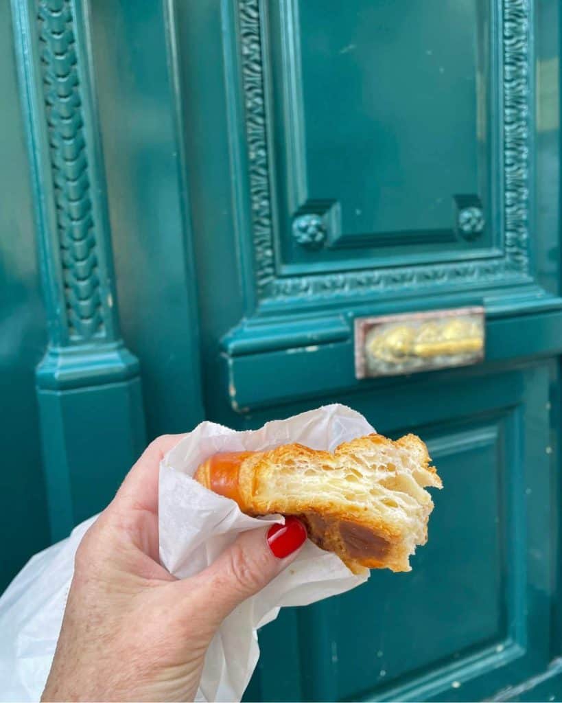 A woman's hand hold a croissant in front of an old teal-colored door in Paris.