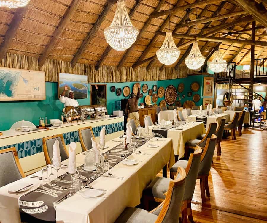 The dining area at Leroo La Tau lodge. A row of tables is set for dinner in a thatched room room with pretty chandeliers and rustic accents.