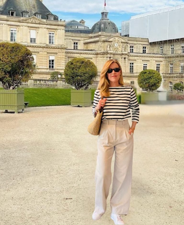 A woman stands in a Paris garden in a striped top, trousers and sneakers. She is holding a straw bag and wearing sunglasses.