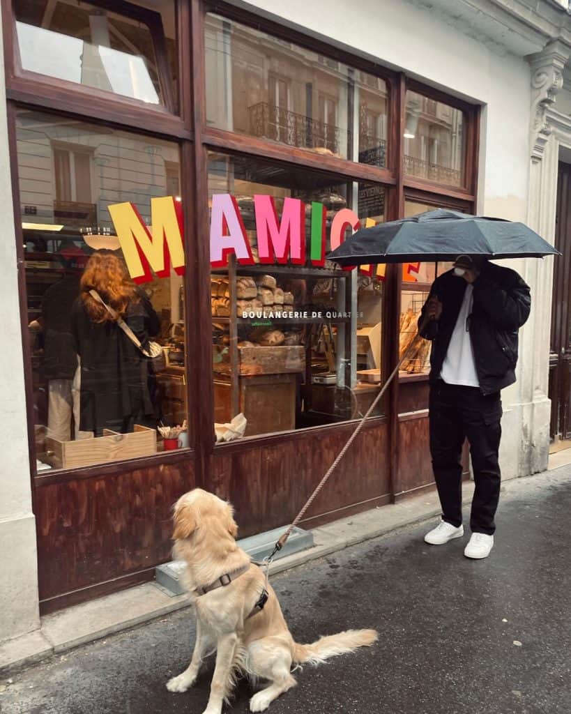 A man stands next to a bakery with an umbrella over his head. He is holding the leash of a golden retriever dog