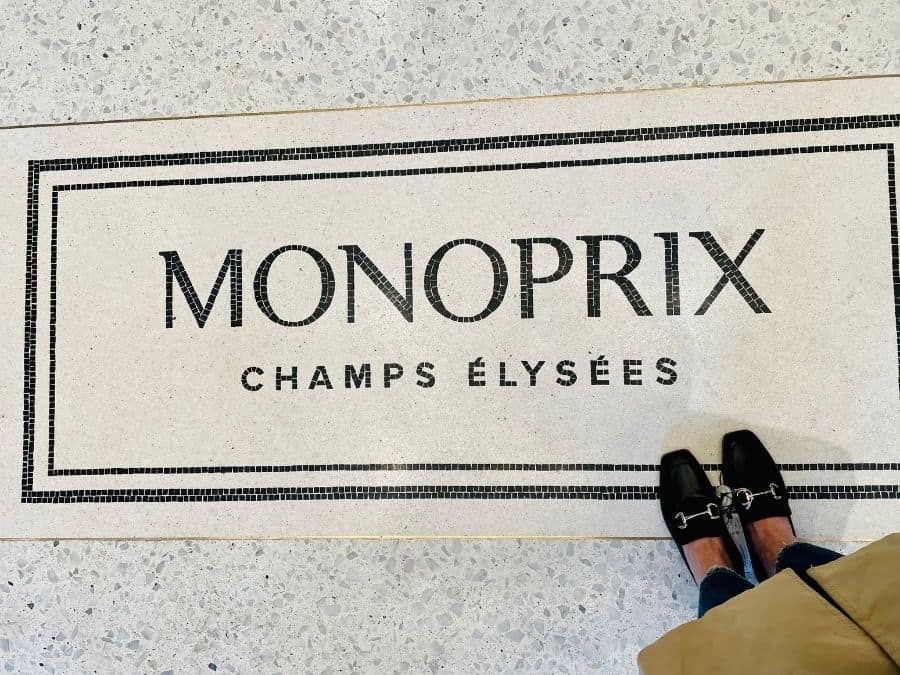 A woman's feet wearing loafers stand on floor tiles with the words Monoprix Champs Elysées, in Paris France.
